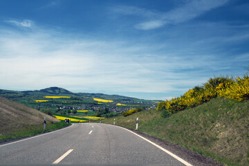 Fahrersicht auf hügelige Landschaft bei Mayen in Rheinland-Pfalz mit Feldern und blühendem Raps - Stockfoto