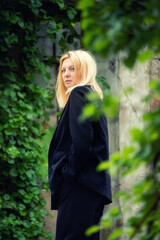 Stylish blonde woman in a black suit standing outdoors near ivy-covered wall. Natural greenery, elegant fashion, and serene expression in a calm urban garden scene.