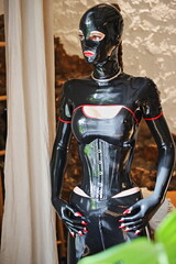 Latex Fetisch Model / Latex BDSM Domina mit schwarzem Hochglanz Latex Outfit, eng geschnürtem Latex Korsett, Latex Maske und langen roten Fingernägeln steht vor einem Fenster