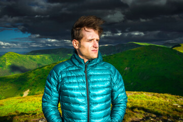 Portrait of a young man in a green down jacket looking away with serious face in windy mountain landscape © XArt