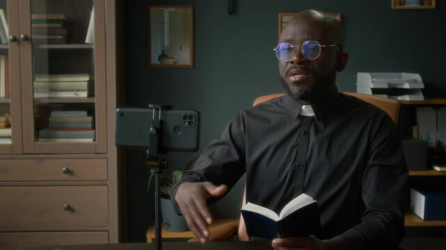 Black young priest wearing glasses having video chat with parishioner providing spiritual guidance to them using smartphone put on tripod