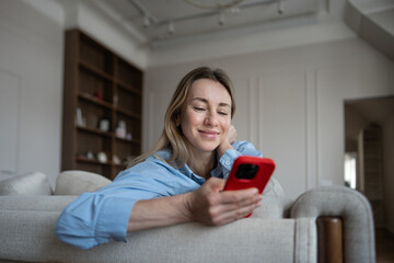 Happy midlife woman lounging on cozy couch at home, scrolling through phone with joy. Relaxed interested mature female smiling reading something pleasant on smartphone resting comfortably in apartment