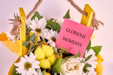 A vibrant flower arrangement features white daisies, yellow decorations, and a pink note reading glorious moments. This cheerful display brightens up any gathering or celebration