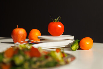 Weighing a fresh tomato on a kitchen scale with fruits and vegetables nearby. Healthy eating and precision in food preparation.