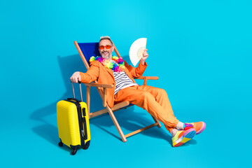 Charismatic middle aged man in orange suit sitting on lounge chair with suitcase and fan on vibrant blue backdrop
