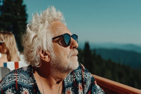 A stylish older man with sunglasses enjoys the sunshine on a balcony with scenic mountain views in the background.