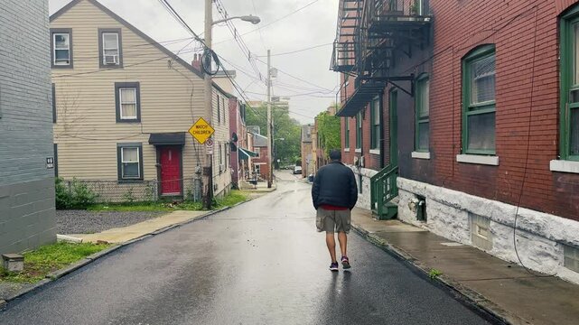 A man walks alone down a quiet Pittsburgh alley on a rainy day. Wet pavement, overcast skies, and historic row houses set the mood in this moody urban scene of solitude and reflection.	