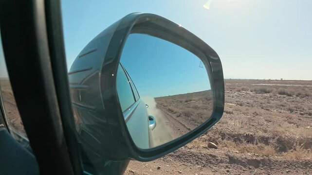 View in rear view mirror from a car driving through a deserted arid terrain. The car is driving on a dusty dirt road. Slow motion 2x