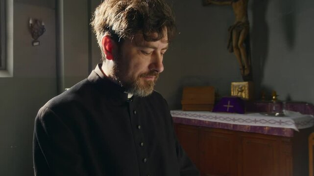 Tilt up side view shot of middle-aged Caucasian clergyman in Catholic soutane standing at table with holy books and golden jug, working on laptop in church with crucifix in background