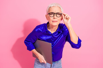 Confident mature businesswoman in blue blouse holding laptop and adjusting glasses on pink...