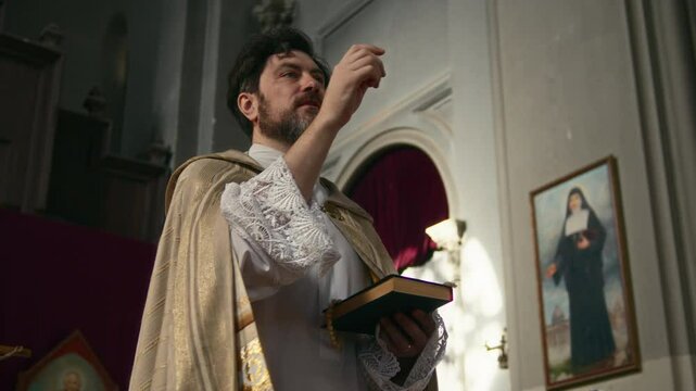 Slowmo shot of middle-aged Caucasian clergyman in golden vestment holding Holy Bible in hands while preaching sermon in Catholic church with icons on walls