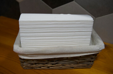 Stack of white paper napkins neatly arranged in a woven basket on a wooden table, showcasing a clean and organized dining setting with a modern aesthetic