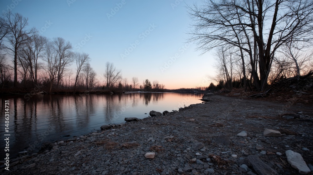 Aufkleber A calm lake captures soft pink light from the setting sun, surrounded by bare trees and a rocky shoreline under a fading sky