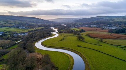 Fototapeta premium Aerial view captures the graceful curve of a river meandering through vibrant farmland and quaint towns, framed by lush hills and forests under a cloudy sky