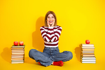 Cheerful young woman in casual striped attire posing on yellow background surrounded by book and apples, promoting education and positivity