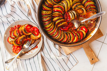 Homemade ratatouille preparation and serving. Child's hands placing vegetable slices into a baking dish. Fresh ingredients, vintage cutlery, cozy kitchen scene, flat lay and side views