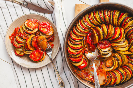 Homemade ratatouille preparation and serving. Child's hands placing vegetable slices into a baking dish. Fresh ingredients, vintage cutlery, cozy kitchen scene, flat lay and side views