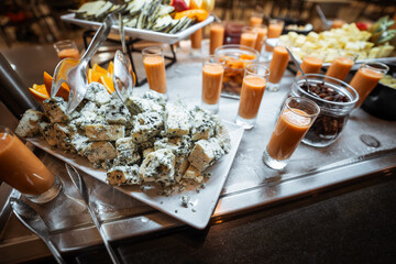 Gourmet buffet table with a close-up of creamy blue cheese cubes garnished with orange slices, surrounded by small glasses of fresh smoothie, dried fruit, and assorted cheese selections