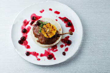Gourmet duck leg with grilled orange, cherry sauce, and creamy risotto on a white plate, and white wooden table