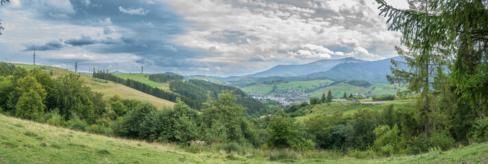 Panoramic view of the Carpathian Mountains with a small town nestled in green hills under a dramatic cloudy sky. Peaceful rural landscape of Western Ukraine.