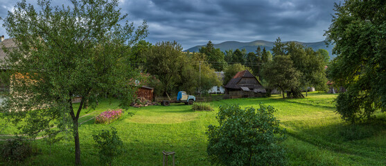 Traditional Carpathian village with wooden houses, lush greenery, and a vintage truck under dramatic skies. Peaceful rural scene nestled in the Ukrainian Carpathian Mountains.