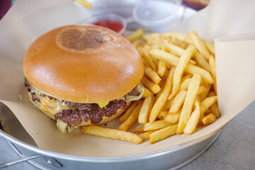 Delicious classic cheeseburger with a juicy beef patty and melted cheese, served with a generous portion of crispy golden french fries on a metal tray. A perfect fast food meal for lunch or dinner.