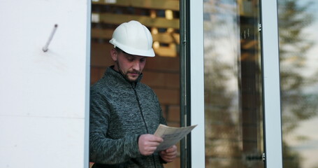 Construction worker studying blueprints in doorway, hard hat on, focused on details of unfinished building project, conveying precision and commitment in construction