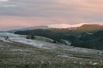 Serene sunrise over snow-dusted mountain pastures. Rolling hills, soft pastel sky, and distant fog create a peaceful landscape perfect for nature lovers and travel enthusiasts.