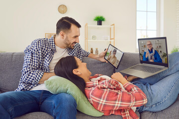Joyful young married couple uses various gadgets to call their elderly parents online. Smiling Caucasian woman and man at home on sofa using laptop and digital tablet for online chat with family.
