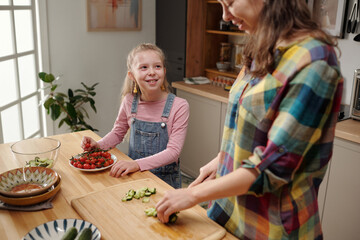 Smiling young girl helping mother by cutting vegetables on wooden countertops in cozy kitchen with natural light coming in from large window