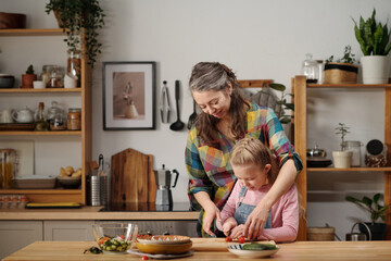 Mother and daughter preparing meal in cozy kitchen with wooden shelves and green plants. Mother embracing daughter while assisting in slicing vegetables on cutting board