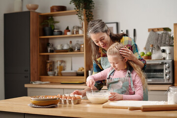 Elderly woman and young girl preparing dough while baking together in modern kitchen with wooden cabinets. Various kitchen tools and ingredients visible on wooden countertop