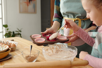 Parent and child baking cupcakes in home kitchen, holding muffin tray with batters next to mixing...