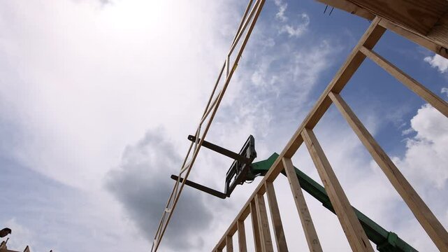 Workers use telehandler lift to position wooden truss in building under construction as clouds gather above.