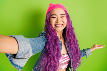 Cheerful young woman with violet hair and casual outfit posing against green background, exhibiting...
