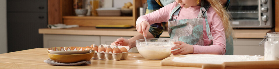 Young girl whisking ingredients in bowl, with support of an adult, while preparing pastries on...
