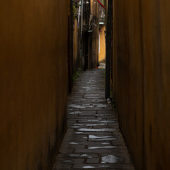 A very narrow and curved street with shabby yellow walls. There are no people on the street. There...