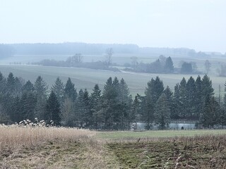 Winter landscape in February without snow Germany Hesse Europe. Idyllic rural landscape with a green hill, framed by bare tree branches under blue sky with clouds. A serene winter countryside scene