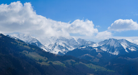Snow-covered mountain range under a bright blue sky with puffy clouds. Spring alpine landscape with green valleys and majestic peaks, perfect for nature and travel themes