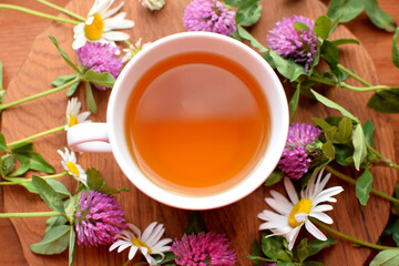 Chamomile medicinal tea in white cup on wooden table with chamomile and red clover flowers. Top view.