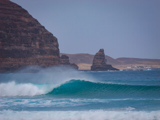 Perfect empty wave rolling beneath rugged volcanic cliffside with sea spray rising into the air....