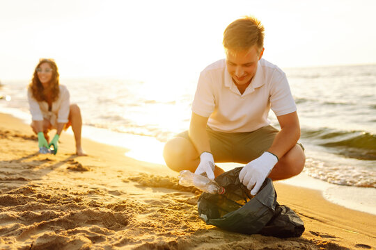 Young friends actively participate in beach cleanup. Cheerful volunteers collect plastic waste and trash along the coastline at sunset. The concept of ecology, ecology and recreation.