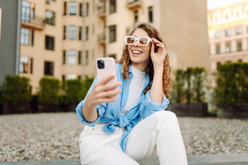Stylish woman in sunglasses taking selfie on sunny city streets. Young woman having fun and blogging at sunset. Blogging, tourism concept.