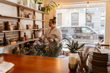 A young White female cafe owner in a white blouse organizing brown paper bags and jars on wooden shelves behind the counter of her bright modern cafe with large windows and indoor plants.