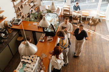 A group of white adult men and women are inside a modern cafe. Some stand at the counter talking to staff, while one man sits by the window using a tablet.
