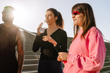 A White adult woman with a ponytail is drinking water from a plastic bottle while standing between a White adult woman in sunglasses and a Black adult man in sunglasses on the stairs