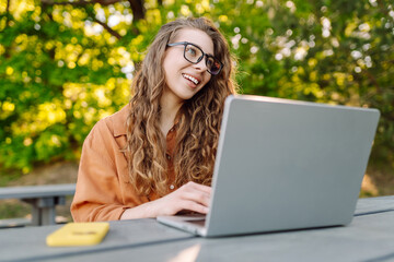 Portrait of young woman in glasses working on laptop in sunny park. Female freelancer at wooden table with laptop and phone outdoors. Remote work concept, technology.
