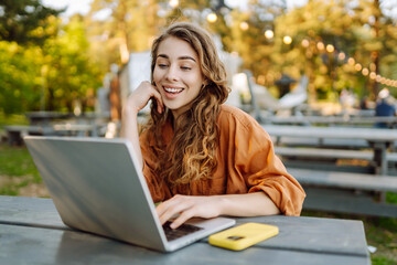 Portrait of young woman in glasses working on laptop in sunny park. Female freelancer at wooden table with laptop and phone outdoors. Remote work concept, technology.
