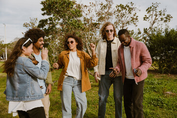 A group of multiracial adult men and women in their 20s and 30s dance and smile together outdoors in a park. They wear casual clothing and enjoy time among friends on a grassy field.