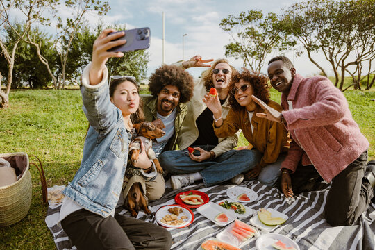 The group of five adult friends sit on a blanket in a sunny park, taking selfies with a dog and sharing fruit during a picnic. They are smiling, eating, and enjoying an outdoor moment together.